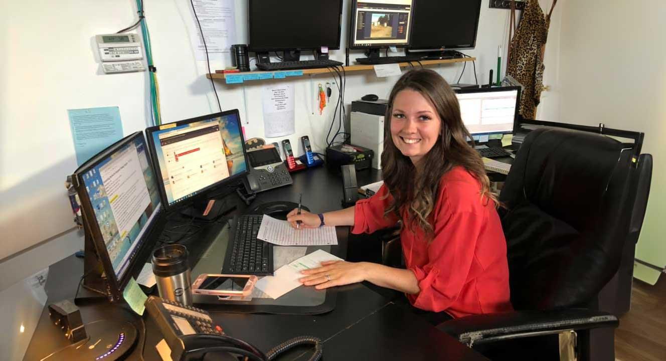 female dispatcher looking toward camera sitting at desk with two monitors