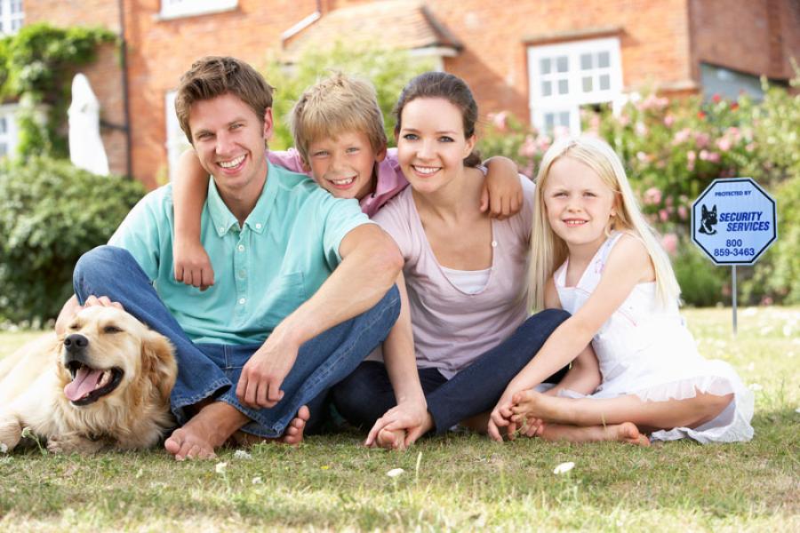 family of four and dog sitting on grass in front of house with alarm company sign in background