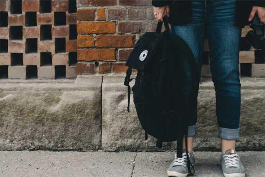 student standing on sidewalk holding backpack, only the legs are visible