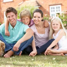 family of four and dog sitting on grass in front of house with alarm company sign in background