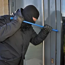 man wearing black clothes and mask using crowbar to break into building