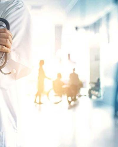 doctor in white coat holding stethoscope, image cropped from shoulders down, standing in hospital hallway