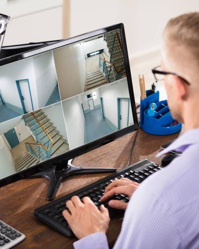 man sitting at desk watching four security camera views on computer screen